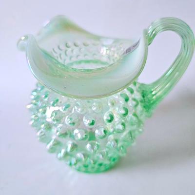Sale Photo Thumbnail #585: This original green iridescent Fenton jug has its Fenton sticker which is silver and says handmade in USA. This hobnail jug has the Fenton stamp on the bottom. It is iridescent, hob nail and stands a total of 5 inches tall with a ruffled top. It is a beau