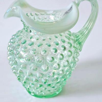 Sale Photo Thumbnail #588: This original green iridescent Fenton jug has its Fenton sticker which is silver and says handmade in USA. This hobnail jug has the Fenton stamp on the bottom. It is iridescent, hob nail and stands a total of 5 inches tall with a ruffled top. It is a beau