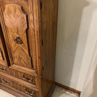 Sale Photo Thumbnail #13: Wooden armoire featuring two paneled doors with carved detailing and decorative brass pulls, opening to reveal shelves. Below are two drawers with brass handles for additional storage. The cabinet shows a natural wood finish and sits on a sturdy base. In 