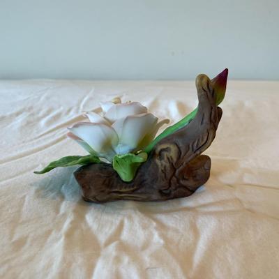 Sale Photo Thumbnail #178: Porcelain rose figurine featuring a detailed pink flower with green leaves, mounted on a wood-like textured base. The piece measures 5 inches in length. It has protective green felt pads on the underside for surface protection. Condition appears good.