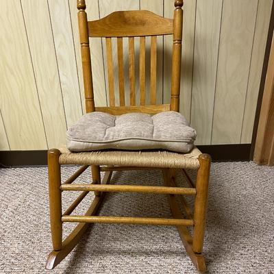 Sale Photo Thumbnail #314: Wooden rocking chair featuring a woven seat and a slatted backrest design. The chair includes a beige cushion for added comfort, securely fastened. The rocking chair measures approximately 33.5 inches high and has a 17-inch width. The wooden frame showcas