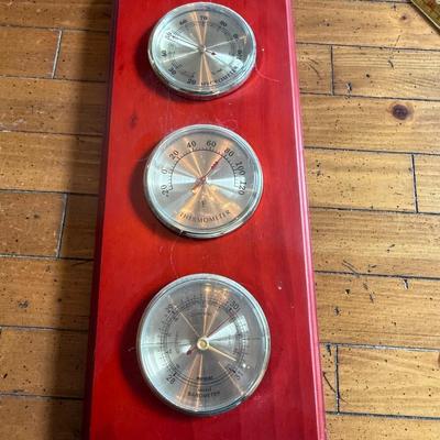 Sale Photo Thumbnail #254: Description: Wall-mounted weather station with three brass-tone dials — hygrometer, thermometer, and barometer — on a red-stained wood base. Vintage style.