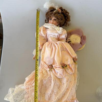 Sale Photo Thumbnail #177: Elegant vintage-style porcelain doll in a peach satin and lace gown, detailed with rose accents and a feathered hairpiece. Includes matching hat.