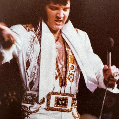 Sale Photo Thumbnail #524: The image shows Elvis Presley performing in concert. Based on the costume he is wearing, particularly the white jumpsuit with the eagle design, it is likely that this image was taken during his 1973 "Aloha From Hawaii" special, or a subsequent performance