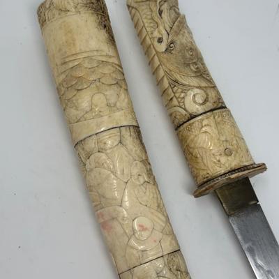 Sale Photo Thumbnail #73: 9 3/4” blade and 20” overall
19th century Japanese Meiji Era tanto samurai knife with a straight single-edged blade. It has ivory or ox bone hand carved mounts depicting traditional scenes. The sheath is chipped at the bottom.