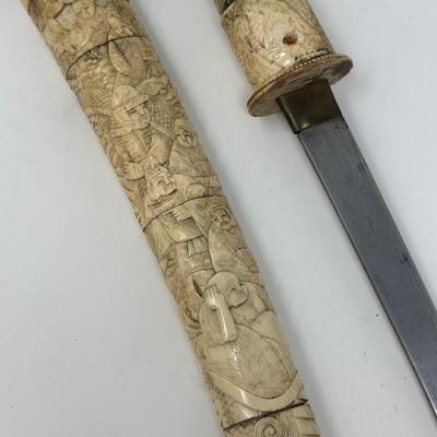 Sale Photo Thumbnail #70: 9 3/4” blade and 20” overall
19th century Japanese Meiji Era tanto samurai knife with a straight single-edged blade. It has ivory or ox bone hand carved mounts depicting traditional scenes. The sheath is chipped at the bottom.