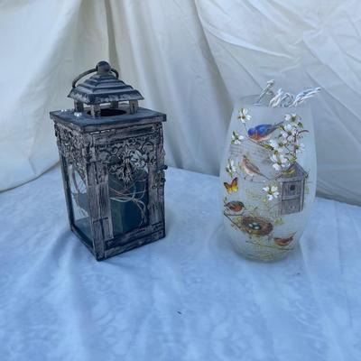 Sale Photo Thumbnail #87: Two decorative lamps, each approximately 8" tall. Left lantern is made of metal with glass sides, has a latched door and a ring on top for hanging, and has battery operated string lights and fake foliage inside. Right lamp is a glass vase with bird graphi