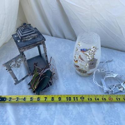 Sale Photo Thumbnail #89: Two decorative lamps, each approximately 8" tall. Left lantern is made of metal with glass sides, has a latched door and a ring on top for hanging, and has battery operated string lights and fake foliage inside. Right lamp is a glass vase with bird graphi