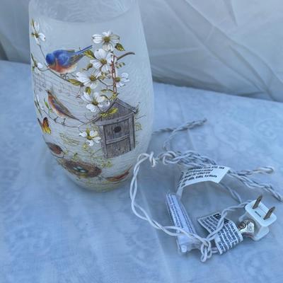 Sale Photo Thumbnail #86: Two decorative lamps, each approximately 8" tall. Left lantern is made of metal with glass sides, has a latched door and a ring on top for hanging, and has battery operated string lights and fake foliage inside. Right lamp is a glass vase with bird graphi