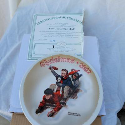 Sale Photo Thumbnail #204: This is a Norman Rockwell collector plate titled "The Christmas Sled," dated 2002. 
Artist:
Norman Rockwell, a renowned American painter known for his heartwarming depictions of American life. 
Theme:
Christmas, featuring a scene of children and a dog sle