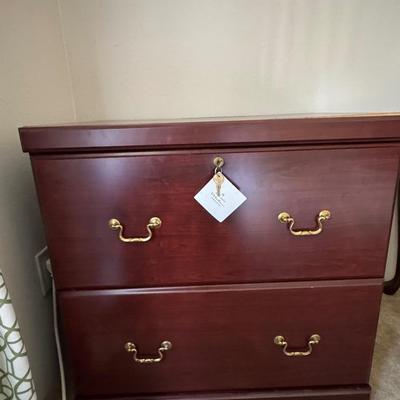 Wooden, two-drawer filing cabinet with brass-toned handles and a key in the top drawer's lock.