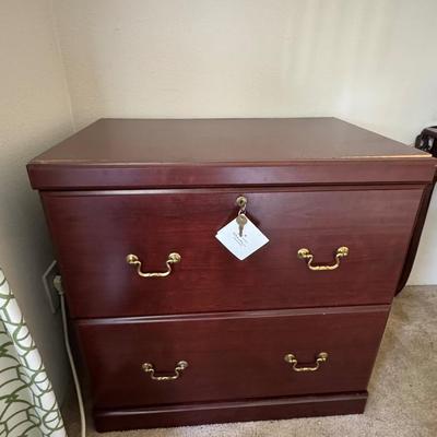 Wooden, two-drawer filing cabinet with brass-toned handles and a key in the top drawer's lock.