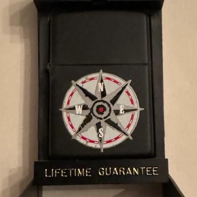 Sale Photo Thumbnail #209: Zippo lighter with a compass rose design on a black matte finish. The text on the lighter indicates it is a Zippo brand lighter, made in the U.S.A., with a lifetime guarantee. It appears to be a Marlboro Compass Rose Zippo lighter, likely from 1998,