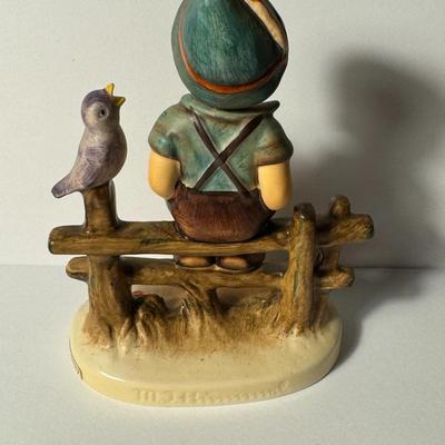 Sale Photo Thumbnail #309: Hummel figurine titled "Wayside Harmony". It is model number 111. The figurine depicts a young boy dressed in traditional lederhosen and a hat, sitting on a fence while being entertained by a chirping songbird perched on one of the posts. This particular 