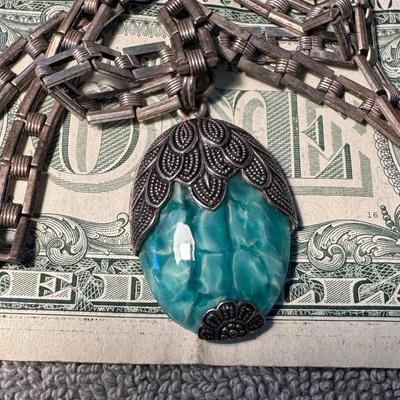 Sale Photo Thumbnail #508: necklace featuring a distinctive pendant and chain:
Pendant: The large, central pendant is made of a translucent, turquoise-colored material, possibly resin or a type of stone like chrysocolla or dyed howlite, with a unique, marbled or fractured appearanc