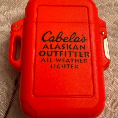 Sale Photo Thumbnail #223: Cabela's Alaskan Outfitter All-Weather Lighter . It is an outdoor lighter designed to be reliable in various weather conditions. 
The lighter's characteristics include:
Brand: Cabela's 
Model/Series: Alaskan Outfitter, related to the Alaskan Guide series,