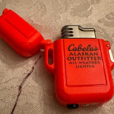 Sale Photo Thumbnail #224: Cabela's Alaskan Outfitter All-Weather Lighter . It is an outdoor lighter designed to be reliable in various weather conditions. 
The lighter's characteristics include:
Brand: Cabela's 
Model/Series: Alaskan Outfitter, related to the Alaskan Guide series,