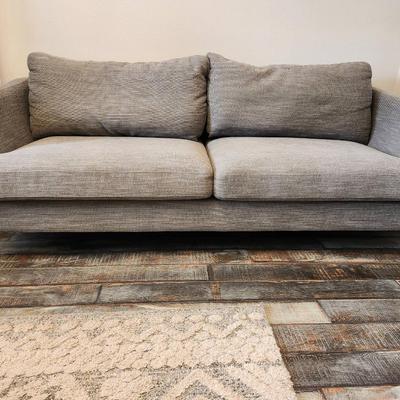 Sale Photo Thumbnail #2: Gray upholstered sofa featuring a textured fabric design and two large seat cushions. Includes a pair of matching backrest cushions for added comfort. Supported by four angled, mid-century modern style wood legs. Dimensions not specified.
