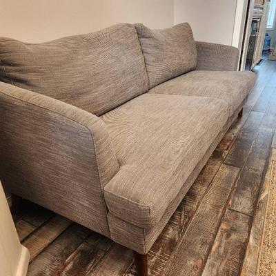 Sale Photo Thumbnail #4: Gray upholstered sofa featuring a textured fabric design and two large seat cushions. Includes a pair of matching backrest cushions for added comfort. Supported by four angled, mid-century modern style wood legs. Dimensions not specified.