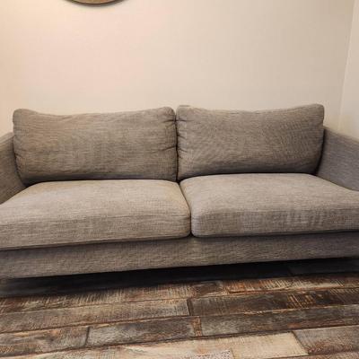 Sale Photo Thumbnail #1: Gray upholstered sofa featuring a textured fabric design and two large seat cushions. Includes a pair of matching backrest cushions for added comfort. Supported by four angled, mid-century modern style wood legs. Dimensions not specified.