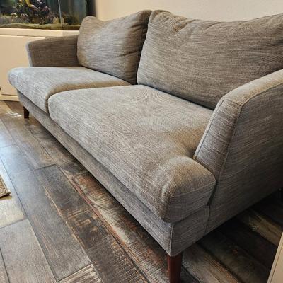Sale Photo Thumbnail #3: Gray upholstered sofa featuring a textured fabric design and two large seat cushions. Includes a pair of matching backrest cushions for added comfort. Supported by four angled, mid-century modern style wood legs. Dimensions not specified.