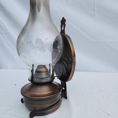 Sale Photo Thumbnail #84: Oil lamp featuring a clear glass chimney supported by a brass base. Includes a reflective metal backing, likely for light amplification. Shows signs of use, including some wear and patina on the brass and minor discoloration on the glass. Supported by thr