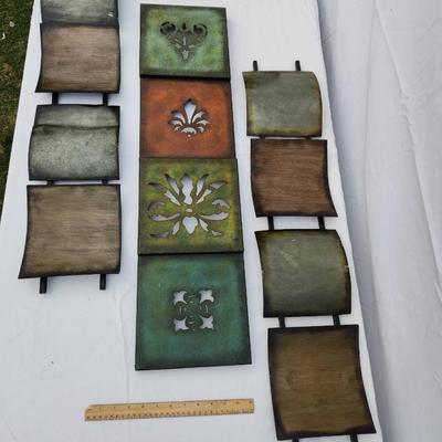 Sale Photo Thumbnail #160: Nine pieces of metal wall decor featuring square panels with various finishes, including green, brown, and rust tones. The central panels have decorative cutout designs: a fleur-de-lis, a floral motif, and a cross shape. The surrounding panels are texture