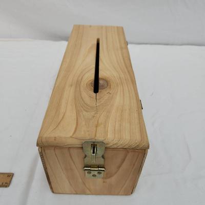 Sale Photo Thumbnail #178: Rectangular wooden storage box with a hinged lid and a narrow slot on the top. The box is equipped with a metal latch at one end for secure closure. Made from natural wood with visible grain and knots, the box allows for potential insertion of objects thr