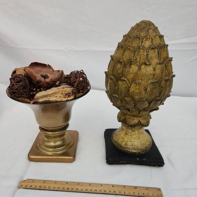 Sale Photo Thumbnail #186: Decorative home decor items. Includes a gold-tone urn with flared bowl and potpourri, featuring dried botanical elements such as wood chips and woven spheres. Also includes a gold-tone textured artichoke-shaped finial on a black base. Both items show wear