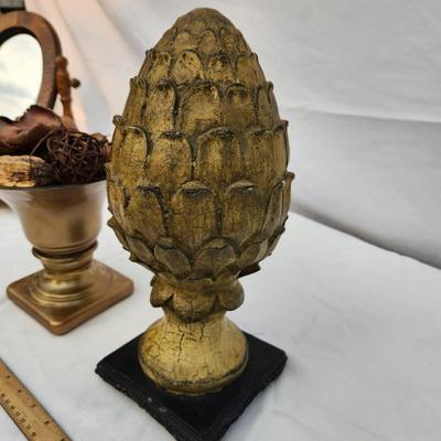 Sale Photo Thumbnail #187: Decorative home decor items. Includes a gold-tone urn with flared bowl and potpourri, featuring dried botanical elements such as wood chips and woven spheres. Also includes a gold-tone textured artichoke-shaped finial on a black base. Both items show wear