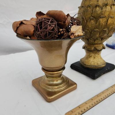 Sale Photo Thumbnail #188: Decorative home decor items. Includes a gold-tone urn with flared bowl and potpourri, featuring dried botanical elements such as wood chips and woven spheres. Also includes a gold-tone textured artichoke-shaped finial on a black base. Both items show wear