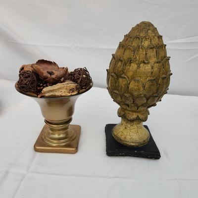 Sale Photo Thumbnail #185: Decorative home decor items. Includes a gold-tone urn with flared bowl and potpourri, featuring dried botanical elements such as wood chips and woven spheres. Also includes a gold-tone textured artichoke-shaped finial on a black base. Both items show wear