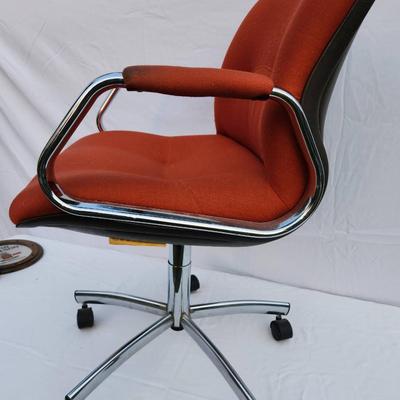 Sale Photo Thumbnail #216: Mid-Century Modern office chair featuring an orange upholstered seat and back, complemented by a chrome metal frame and armrests for a sleek design. The chair is equipped with a five-star chrome base and caster wheels for mobility and an adjustable height