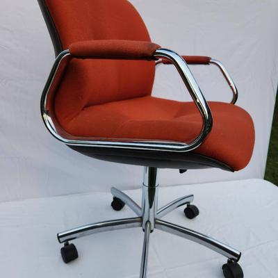 Sale Photo Thumbnail #215: Mid-Century Modern office chair featuring an orange upholstered seat and back, complemented by a chrome metal frame and armrests for a sleek design. The chair is equipped with a five-star chrome base and caster wheels for mobility and an adjustable height