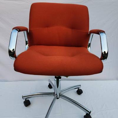 Sale Photo Thumbnail #214: Mid-Century Modern office chair featuring an orange upholstered seat and back, complemented by a chrome metal frame and armrests for a sleek design. The chair is equipped with a five-star chrome base and caster wheels for mobility and an adjustable height