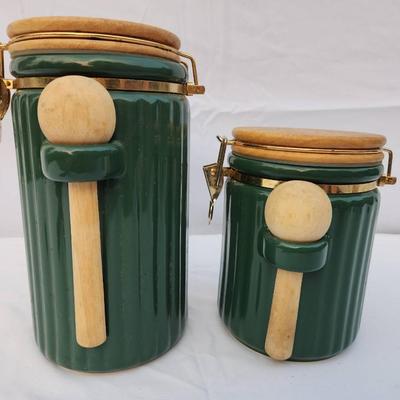 Sale Photo Thumbnail #240: Two ribbed ceramic canisters in a green finish, featuring hinged wooden lids and matching wooden spoons secured on the fronts. Each canister is fitted with a clasp closure to ensure a tight seal. These functional and decorative items are suitable for hold