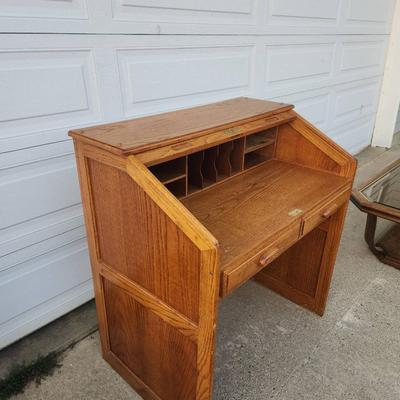 Sale Photo Thumbnail #299: Oak roll-top desk featuring a tambour front that slides open to reveal storage compartments and cubbyholes. Includes two pull-out drawers beneath the desktop for additional storage. The desk showcases a natural wood grain finish and paneled sides. Ideal f