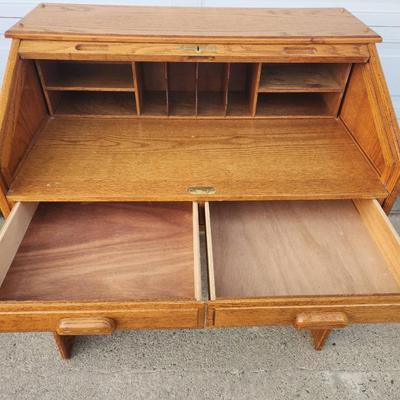 Sale Photo Thumbnail #298: Oak roll-top desk featuring a tambour front that slides open to reveal storage compartments and cubbyholes. Includes two pull-out drawers beneath the desktop for additional storage. The desk showcases a natural wood grain finish and paneled sides. Ideal f