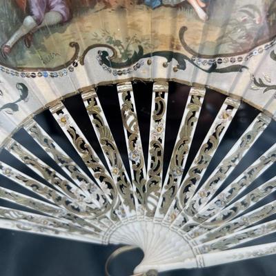 Sale Photo Thumbnail #397: A folding hand fan, antique, in a shadow box frame   Mother of pearl on the  sticks with the scene likely painted on silk or similar material. The sticks are likely bone, carved and pierced.  Signed by Lucien. Shadow box is 20 3/4 x 12 inches.  
Retails a