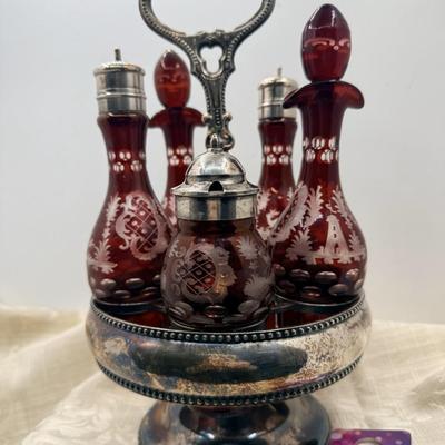 Sale Photo Thumbnail #422: From late 19th or early 20th Century, 5 ruby red glass bottles with 'cut to clear' etchings of trees, buildings, and deer.  
Held in silver plate stand, which needs polishing.  Bottles are for oil, vinegar, salt, pepper, and mustard.
13 inches tall with s