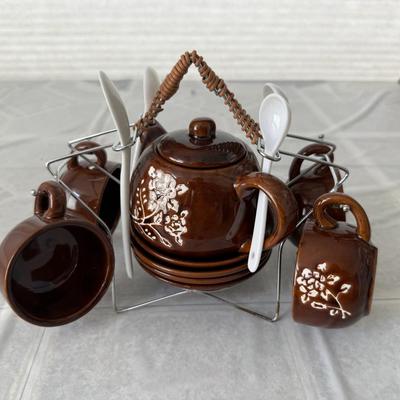 Sale Photo Thumbnail #9: The set is made of brown ceramic or redware pottery with a glossy finish and hand-painted floral details.
It includes a teapot, four cups, four saucers, and four spoons, all held in a wire metal caddy with a woven handle.