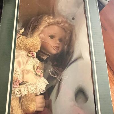 Sale Photo Thumbnail #15: Charming Geppeddo porcelain doll with blonde curls, lace-trimmed dress in rose pink, and small teddy bear accessory. Comes in original green display box with viewing window. Doll appears to have never been removed from box.
Box shows some creasing and sto