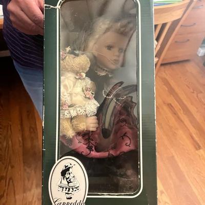 Sale Photo Thumbnail #13: Charming Geppeddo porcelain doll with blonde curls, lace-trimmed dress in rose pink, and small teddy bear accessory. Comes in original green display box with viewing window. Doll appears to have never been removed from box.
Box shows some creasing and sto