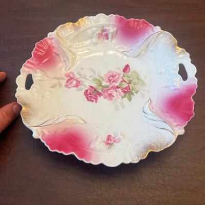 Sale Photo Thumbnail #57: Delicate antique German porcelain plate featuring a hand-painted pink rose motif, scalloped edge, and gold detailing. Embossed with elegant reliefs and dual handles, this piece exemplifies early 20th-century craftsmanship.
Details:
•	Maker: Likely RS Ge