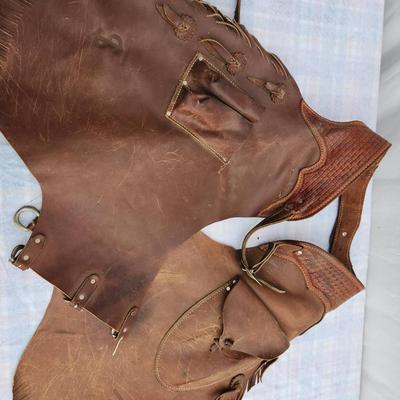 Sale Photo Thumbnail #36: Rugged leather chaps featuring fringe accents and a utility pocket. Adjustable buckle straps ensure a customizable fit. The leather shows visible signs of use and wear, including scuff marks and discoloration, indicating prior practical use. The chaps are