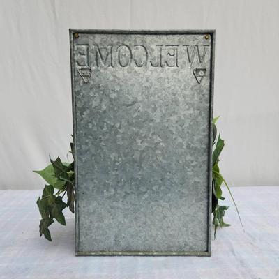 Sale Photo Thumbnail #142: Galvanized metal welcome sign featuring embossed lettering and an attached faux greenery arrangement including ivy and variegated leaves. The sign includes a brass-tone label holder on the front for easy personalization. Suitable for indoor or covered out