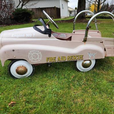 Sale Photo Thumbnail #11: Antique Pink pedal car featuring 'Fire and Rescue' and 'Engine No 7' decals, a metal body, and chrome accents, including handlebars and steering wheel. Designed with a spacious back area and working pedal system. The car shows signs of use, including visi