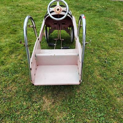 Sale Photo Thumbnail #14: Antique Pink pedal car featuring 'Fire and Rescue' and 'Engine No 7' decals, a metal body, and chrome accents, including handlebars and steering wheel. Designed with a spacious back area and working pedal system. The car shows signs of use, including visi