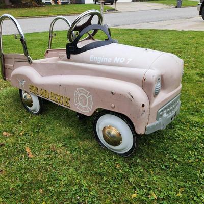 Sale Photo Thumbnail #13: Antique Pink pedal car featuring 'Fire and Rescue' and 'Engine No 7' decals, a metal body, and chrome accents, including handlebars and steering wheel. Designed with a spacious back area and working pedal system. The car shows signs of use, including visi