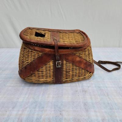 Sale Photo Thumbnail #148: Wicker fishing creel with leather trim and straps, featuring a lid with a slot for fish entry and a buckle closure mechanism for securing the contents. The creel is designed for outdoor fishing use, with carrying straps for convenience. In used condition,